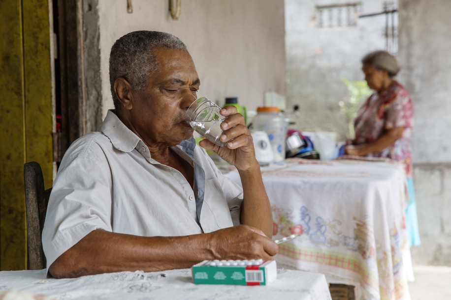 Daniel Lordelo Vaz, aged 77, taking his diabetes medications while his wife Maria Filomena, aged 73, stands in the background. The couple lives with family relatives in Leandrinho, Dias D'Àvila, Bahia, Brazil.