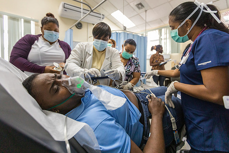 Emergency room nurses and doctors perform a routine process on Kelly, acting as a patient experiencing a severe anaphylactic reaction during the observation period after receiving the vaccine, during a vaccination drill conducted at the Siparia District Health Facility in Trinidad and Tobago on 6 March 2021. As Trinidad and Tobago prepare for their initial delivery of 100,000 to 120,000 doses of the Oxford-AstraZeneca COVID-19 vaccine, a key strategy in preparing and training the healthcare workforce is the use of simulation exercises. This includes the use of different simulation exercises, including discussion based table-top exercises as well as more operational based exercises such as drills. They are used to assist in developing, assessing and testing the functional capabilities of emergency systems, procedures and mechanisms to respond to public health emergencies. COVID-19 vaccine simulations test planning assumptions and procedures in a safe and constructed environment before national vaccine roll-outs occur. These simulations help to ensure that vaccine roll-outs have the best possible chance of success. Using simulations developed by WHO and COVAX, health workers in Trinidad and Tobago are able to test and practice the planning and logistics of vaccine administration within the specific context of their country. Participants in these exercises are personnel who perform their actual responsibilities and respond as they would in a real situation. They can practice their role within the vaccine roll-out at every stage and identify where the plan and procedures are working well and where it could be improved. During the vaccination drill conducted at the Siparia District Health Facility in Trinidad, multiple scenarios were simulated at once to test preparedness including: a recipient who was fully decided to take and receive the vaccine, a recipient who was undecided, walk in recipients with no appointments, a recipient with severe reaction, and a recipient who declined being vaccinated.